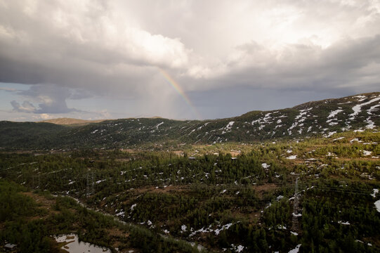 Aerial view of a vibrant rainbow arches over the undulating hills and verdant forests dotted with melting snow under a dramatic sky, Dalen, Telemark, Norway.