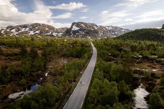 Aerial view of two people walking on a winding road cuts through lush green forests, leading towards majestic mountains capped with remnants of snow, Dalen, Telemark, Norway.