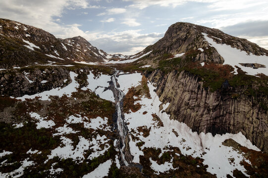 Aerial view of rugged mountain peaks dusted with snow embrace a cascading waterfall, reflecting the sky's soft light, Dalen, Telemark, Norway.