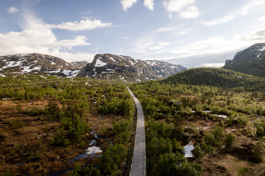 Aerial view of a narrow road cutting through a landscape of green trees, brown fields and snow-capped mountains under a partly cloudy sky, Dalen, Telemark, Norway.