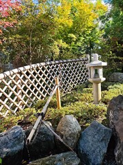 A serene Japanese-style garden scene with a traditional bamboo fountain (shishi-odoshi), a stone lantern and a lattice bamboo fence, set among lush greenery and autumnal foliage.