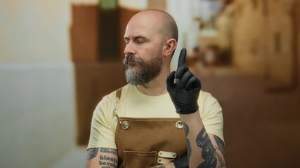 Man with beard and tattoos wearing apron and gloves pointing in urban outdoor setting with closed eyes showcasing focus and determination on city street.