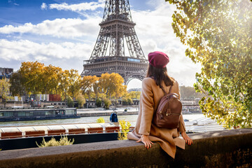 A tourist woman with a beret hat sits at the riverbank of the Seine and enjoys the beautiful view of the Eiffel Tower in Paris, France © moofushi