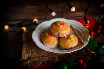 A warm and festive still life of Christmas-themed cupcakes topped with colorful candied fruit. The cupcakes are arranged with holiday decorations including pine branches, red berries, and rustic.