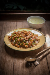 Delicious Thai-style fried chicken with rice, topped with crispy garlic and herbs, served with a bowl of clear soup on a wooden table. Authentic homemade Asian meal.