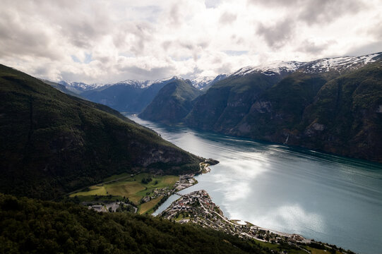 Aerial view of the fjord cutting through the green valleys and towering snow-dusted mountains under a sky of shifting clouds, Aurland, Vestland, Norway.