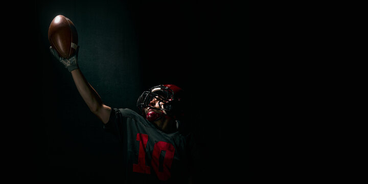 Football player raising ball upward with dramatic emotional expression on dark background. Concept of motivational posters, advertising, branding, hero banners, inspiring sport materials. Banner