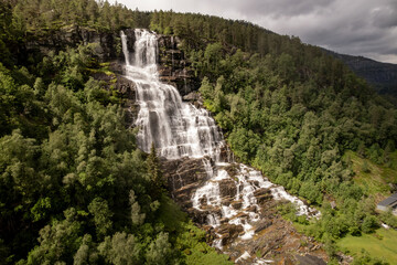 Aerial view of the cascading Tvindefossen waterfall plunges down the rocky cliffside amidst lush green trees, Skulestadmo, Vestland, Norway.