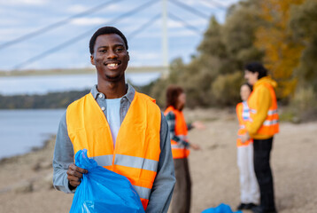Portrait of smiling male volunteer in a safety vest holding trash bags during a community environmental cleanup event.