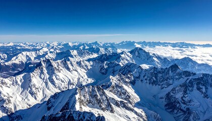 Aerial view of snow-capped mountains under a clear blue sky, with clouds filling some valleys