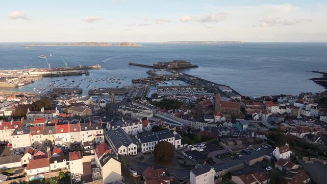 High circling flight over rooftops of St Peter Port Guernsey centring on harbour and Castle Cornet with Herm Sark and Jethou in the distance in bright afternoon sunshine on calm day