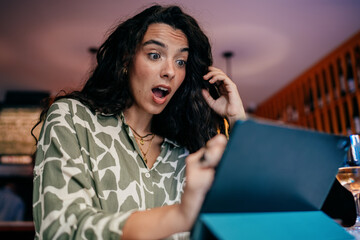 Surprised young woman reacts with wide eyes while holding stylus near tablet, fully immersed in a...