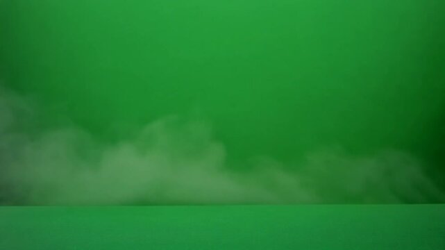 White fog or smoke hovering close to the ground over a uniform green background and floor, creating a dramatic and atmospheric scene suggestive of a green screen setup.