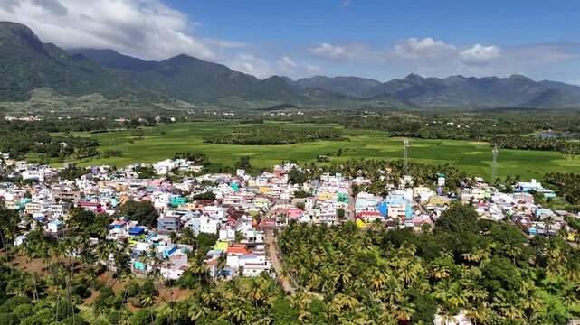 Sweeping drone footage of an expansive town Courtallam Tamil Nadu, set across a hilly landscape. The settlement is characterized by houses interspersed with large patches of green farmlands.