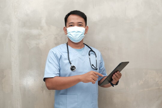 Southeast Asian male healthcare worker in scrubs and face mask using tablet, isolated on light wall, ideal for medical ads or safety campaigns.
