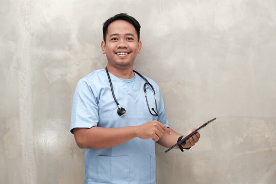 Confident Southeast Asian male nurse or doctor holding a tablet and wearing scrubs with stethoscope, perfect for medical ads and healthcare visuals.

