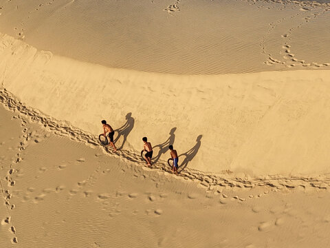Aerial view of three figures casting long shadows across the rippled sands, their footprints etching transient paths in the golden dunes, Ninh Phuoc, Ninh Thuan, Vietnam.