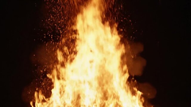 A large fire burns brightly against a black background with sparks flying to the left