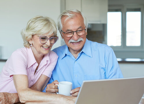 Portrait of a lovely senior mature couple using a laptop together and having fun drinking coffee or tea sitting at a table at home - Powered by Adobe