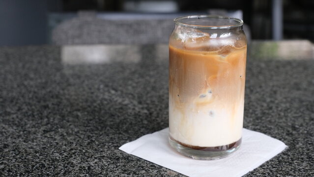 A refreshing iced latte with milk and espresso served in a clear glass jar, placed on a napkin over a granite table surface.