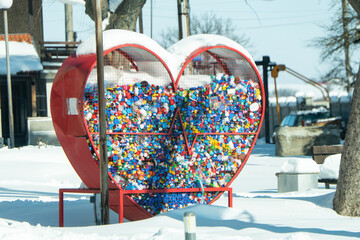 Heart-shaped recycling bin filled with colorful plastic bottle caps, surrounded by snow, showcasing community efforts in environmental sustainability and creative design