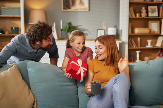 A family enjoys a heartfelt moment in their living room as a child surprises her mother with a gift while the father looks on happily, creating a warm atmosphere.