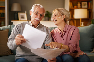 An elderly couple, the man with glasses and a mustache, sit on a sofa at home, reviewing papers, possibly life insurance documents. The woman laughs as she points to a document.
