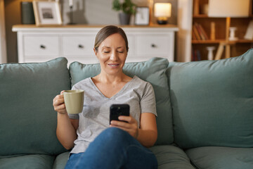 Obraz premium A woman with short hair sits comfortably on a green couch, holding a coffee mug in one hand and checking her smartphone with a smile on her face, enjoying her time in a warm living space.