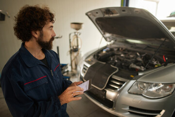 A car mechanic stands beside a silver vehicle with the hood raised, closely inspecting the engine while writing down important information in a notepad.