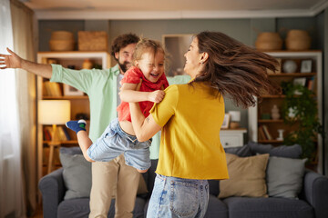 A cheerful family is having fun at home, with laughter filling the air as the mother lifts her daughter while the father joins in the celebration.