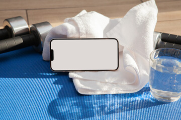Smartphone mockup on towel with dumbbells, glass of water, and yoga mat in sunlight. Home fitness...