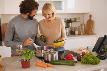 Two people share joyful moments while cooking in a bright kitchen. They prepare fresh vegetables and ingredients, smiling and engaging in each other’s company.
