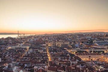 As the sun sets, Lisbon's rooftops glow in warm hues. The river reflects the fading light, while people below enjoy the evening atmosphere, bustling with life