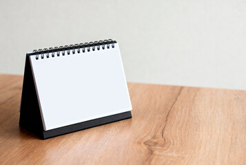 There is a clean calendar layout on the wooden table. The calendar has a spiral binding and a simple black base. Mockup calendar with an empty space for text.