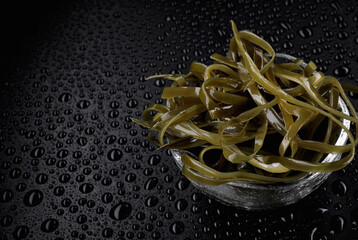 Seaweed salad in a transparent glass cup on a wet black background with water drops. Healthy food.
