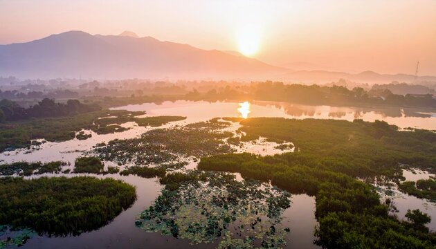 An aerial view captures a serene lake at sunrise, shrouded in mist, with dense green vegetation and mountains in the background.