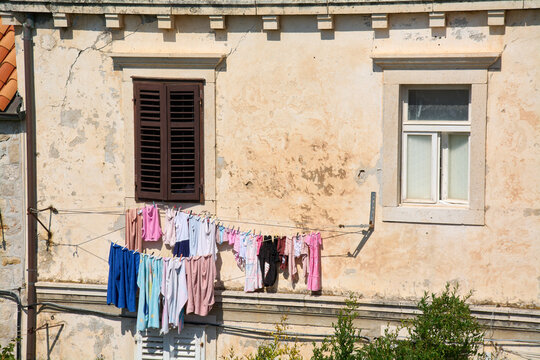 Colorful laundry, including children's clothes, hangs on a clothesline between a brown-shuttered window and a white window on a weathered stucco facade.