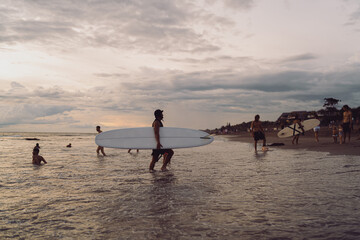 Male surfers wade through shallow ocean water at dusk, carrying long surfboards under a dramatic cloudy sky, representing community, persistence, and connection with natural forces