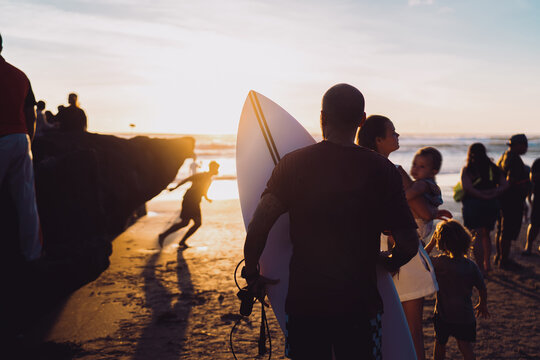 Man holding a surfboard stands on the sand watching the sunset, surrounded by people enjoying the moment. The image conveys freedom, mindfulness, and connection to nature through shared experience