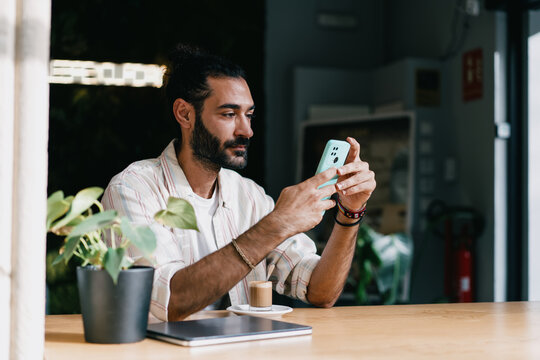 Man smiling while browsing smartphone at café table. Conceptual stock image symbolizing online engagement, digital culture, mobile lifestyle, and authentic personal connection.