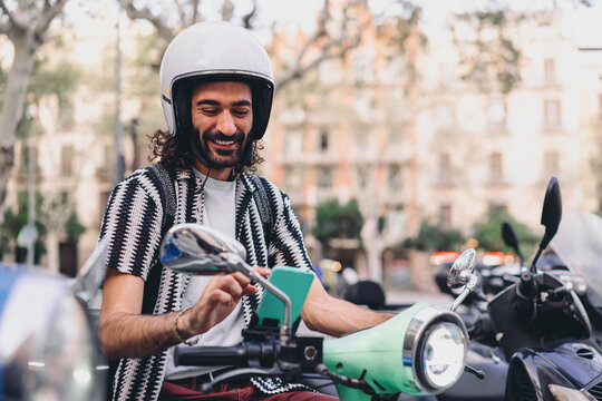 Smiling man sits on scooter using smartphone, surrounded by bikes, enjoying mobile tools and intuitive navigation in modern connected commuting life.