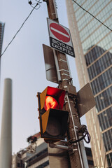 Vertical close-up of a red pedestrian signal and a no-entry sign on a street pole in natural light. Shot from a low angle with tall city buildings in the background.