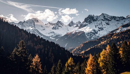 A panoramic view of snow-covered mountain peaks towering over a dense forest displaying autumn foliage in shades of yellow and orange.