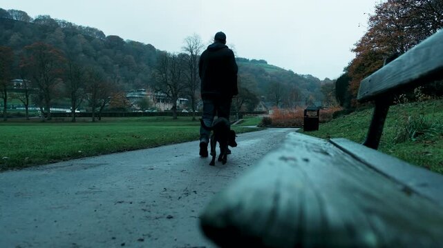 Low angle early morning shot in Todmorden showing a park bench in the foreground leading to a man walking his black lurcher dog on a lead away from the camera