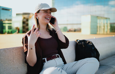 Relaxed young woman sitting cross-legged outdoors with cap, laughing on smartphone, representing...