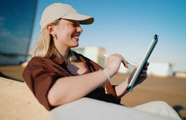 Young woman with cap smiling while drawing on tablet with stylus, representing freelance work, mobile creativity, digital empowerment and independence in technology-driven life. © BullRun