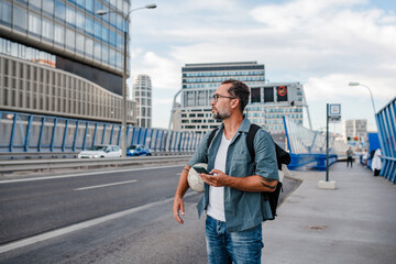 Bearded man with ball under arm using phone while waiting for bus.