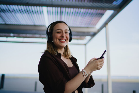 Joyful young woman smiling at camera while holding smartphone and wearing headphones, symbolizing connection, digital lifestyle, entertainment and cheerful mobile experience.