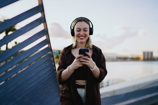 Young woman standing with headphones and smartphone, smiling at screen, symbolizing joyful technology use, digital lifestyle, online connection and cheerful mobile moment. - Powered by Adobe