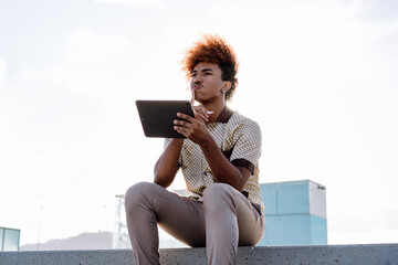Portrait of African American man with curly ginger afro sitting outdoors, holding digital tablet and stylus with finger on lips, showing creative thinking and mobile technology use.